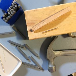 Stämpla text på läderbiten och fyra hål i båda ändorna. Metallstämplar, hammare, nålar, läderbit och träbit på ett bord