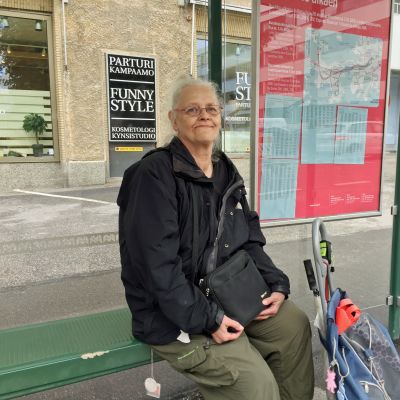 Carita Turunen, en kvinna med vitt hår och glasögon sitter på en busshållplats på Drumsö. Hon har en shoppingkärra bredvid sig och en handväska i famnen.