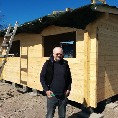 Rakennustöitä vetävä Matti Kinnunen seisoo uuden hirsisaunan edessä aurinkolasit päässä.