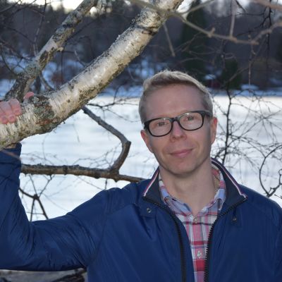 Mies pitää koivun oksasta kiinni rantamaisemassa ja katsoo suoraan kameraan
