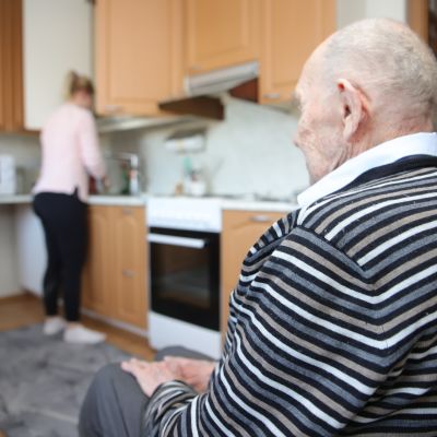 Lähihoitaja tiskaa kotona, 95 vuotias veteraani istuu keittiössä.