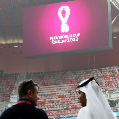 Två personer står inne i en tom arenan, på bildskärmen syns logon för VM 2022.