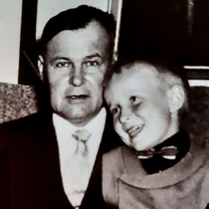 Pieni poika 50-luvun kuvassa istuu onnellisesti hymyillen nojatuolissa istuvan keski-ikäisen miehen sylissä ja nojaa tämän rintaan.