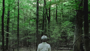 Ryuichi Sakamoto seisoo selin kameraan metsän edessä. Kuva dokumenttielokuvasta Coda.