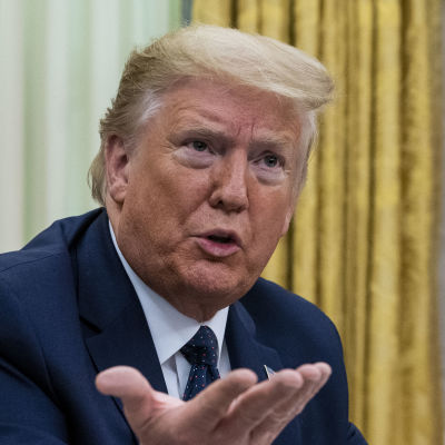 Donald Trump sitter vid ett skrivbord. Han talar och ser arg ut. Han höjer händerna i luften.