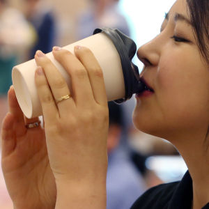 Sydkoreansk kvinna dricker kaffe ur en engångsmugg med ansiktsformat lock.