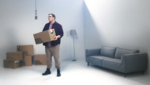 UMK-kilpailija Aksel seisoo vaaleassa huoneessa ja pitää käsissään pahvilaatikkoa, katosta roikkuu vanha mikrofoni ja taustalla lisää laatikoita, jalkalamppu ja sohva.