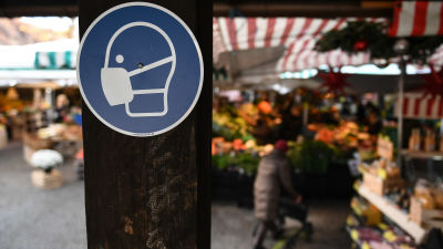 En skylt på en stolpe uppamanar folk att använda munskydd. I bakgrunden ser man en dam titta på varor på en marknad.