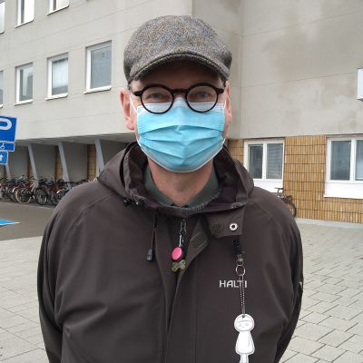 En man med runda svarta glasögon, rutig keps och munskydd står utanför sjukhuset.