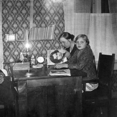  Lampaluodon ensimmäinen radio (kidekone). Radiota kuunnellaan saaristossa pöydän ääressä. Mies, tyttö ja lehteä lukeva nainen.