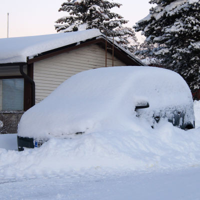 Personbil helt täckt av snö i stor driva på gata i bostadsområde.