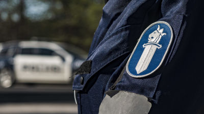Närbild av polisens logo på en polisuniform. I bakgrunden syns en polisbil.