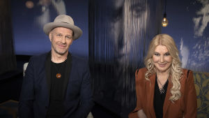 Suurlähettiläistä tuttu laulaja, lauluntekijä Jussu Pöyhönen ja työpsykologian tohtori Helena Åhman kuvatttuna Mediapoliksen studiolla, istuvat vierekkäin, Jussulla hattu päässä.