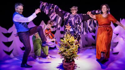 fyra skådespelare dansar kring en liten julgran i ett vinterlandskap på en teaterscen. 