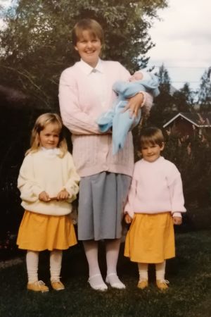 Eivor med sonen Erik i famnen tillsammans med de två äldsta döttrarna Rosmarie och Annsofie. Ett gulnat fotografi av en kvinna. I famnen håller hon ett spädbarn och bredvid henne står två flickar i gula kjolar.