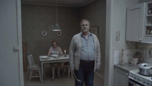 Diskbänksrealism. I Roy Anderssons värld är alla bleka och det mesta grådaskigt. En man står i sitt kök och tittar rakt in i kameran. Hustrun sitter i bakgrunden vid bordet.