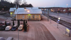 Järnvägsstationen i Karis. En gammal gul järnvägsstation, järnvägsspår, parkerade bilar med isiga vindrutor. Skymning.