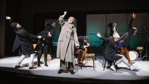 Scen ur Faktiska Händelser på Lilla Teatern. Sex skådespelare i vinterrockar står och sitter på en teaterscen.