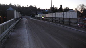 Den nuvarande bron upplevs som otrygg. Den är både smal och i dåligt skick. En bro för bilar och lätt trafik. Halt väglag. Mörk vintermorgon eller -kväll. En cyklist i reflexväst leder sin cykel på den smala trottoaren till höger. Klottrade räcken. Trafikmärke som visar att trafiken i ena riktningen har förkörsrätt.
