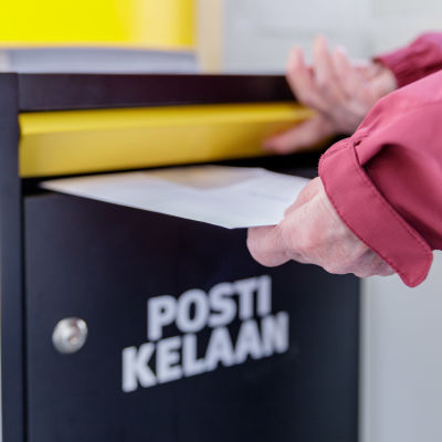 Naisen kädet laittavat kirjeen Kelan postilaatikkoon. Postilaatikossa teksti: Posti Kelaan.