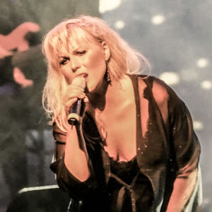 Josefin Nilsson en kvinna med blont hår och svarta kläder står på en scen och sjunger i en mikrofon, i bakgrunden syns en gitarrist.