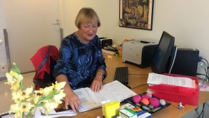Kaarina Tynkkynen har jobbat med missbrukare i decennier. En kvinna i blåbrokig blus sitter och bläddrar i en tidsbokningskalender, på bordet finns dator, skrivare, papper, dekorationer.