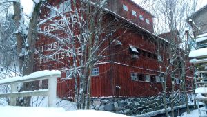 En stor röd gammal träbyggnad vid en liten å. I gaveln står det Fiskars Valiosiementä Elitutsäde. Vinter, snö på träden.