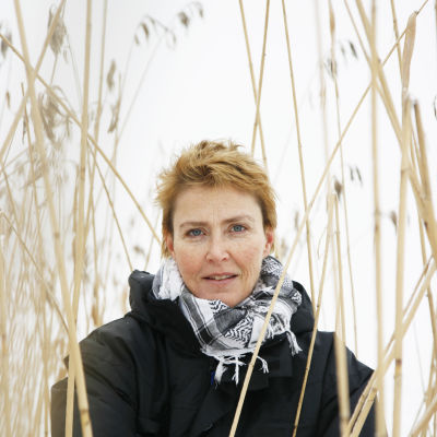 Författaren Minna Lindeberg iklädd en svart jacka och svartvit sjal står ibland vass och tittar in i kameran.