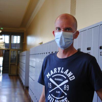 Skallig man med mörkblå t-skjorta med tryck och munskydd står framför skåp i en skolhall.