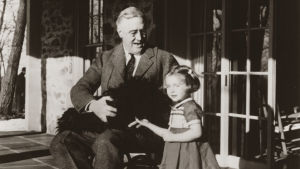 FDR led av polio. Franlin D. Roosevelt som sitter i en rullstol. I famnen har han en hund och bredvid honom står ett barn, en flicka.