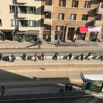 Ihmiset odottavat linja-autoa pitäen runsaasti etäisyyttä toisiinsa.