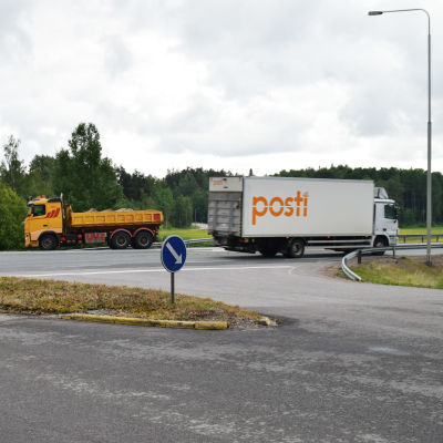 En gul lastbil möter en vit Posti-lastbil i en asfalterad vägkorsning. I förgrunden syns ett stopmärke och i bakgrunden skog och en väg.