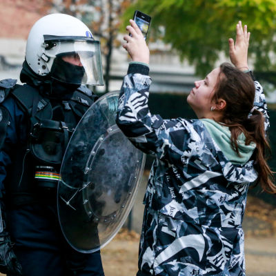 En kvinna som står framför en kravallpolis med sina händer uppsträckta och en telefon i ena handen.  
