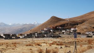 Landsbygden i Tibet har länge hört till de fattigaste delarna av Kina. Nu heter det att absolut fattigdom inte längre existerar heller där. En samling hus på en bergssluttning med vissnat gräs. Snöklädda bergstoppar i bakgrunden.