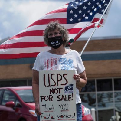 En kvinna med munskydd håller en skylt i handen med texten "USPS är inte till salu". Bakom henne viftar någon med USA:s flagga.
