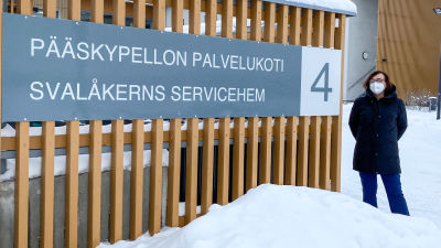 En kvinna i vinterkläder och vitt munskydd står utomhus bredvid ett staket med en stor skylt där det står Svalåkerns servicehem.
