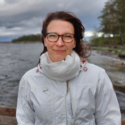 Kvinna med glasögon och mörkt hår står vid en strand och ser mot kameran