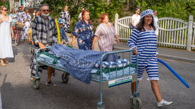 Sjusovartåg längs Appelgrensvägen i Hangö. Soligt. I sängen ligger någon inbäddad och gömd. Folk är klädda i pyjamas.