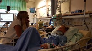 Adams mamma, Mia, hälsade på så ofta hon kunde. adam nyholm ligger i en sjukhussäng med nackkrage och alla möjliga slangar överallt. hans mamma sitter bredvid honom.