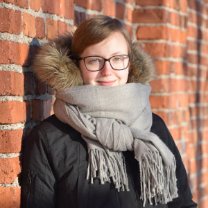 Maria Hagberg. Porträtt på Maria Hagberg, står framför rödtegelvägg i solsken, grå halsduk runt halsen.