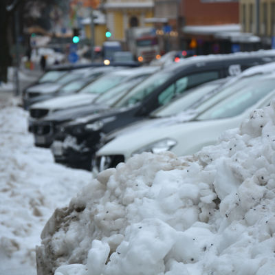 Flera bilar står parkerade i rad framför en stor snöhög. 