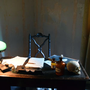 Installation som föreställer prof. Saelans skrivbord Gammalt skrivbord med lampa, apoteksflaskor, anteckningar, bläckpenna.