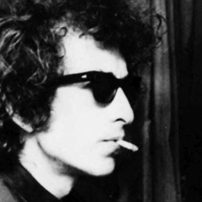 Bob Dylan tupakka suussa ja aurinkolasit silmillään. Taustalla mies kuvaa elokuvakameralla.