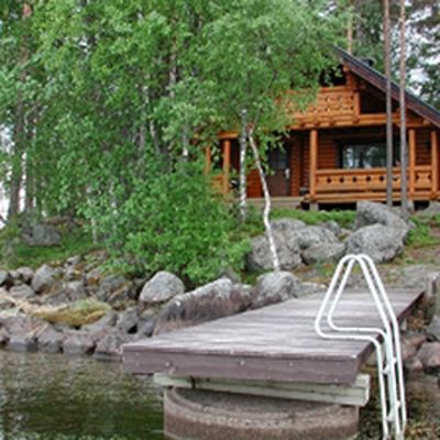 Kuvassa hirsinen huvila kivikkoisella järvenrannalla kesäisessä maisemassa, etualalla laituri.