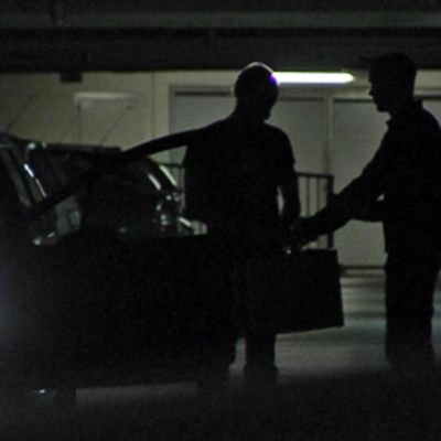 Kaksi ihmistä juttelee keskenään hämärässä parkkihallissa. Oikealla oleva mies ottaa salkun vastaan.