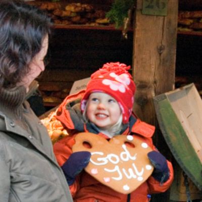 Punapipoinen pikkutyttö pitelee isoa piparkakkua, jossa teksti "God Jul". Toisella puolella äiti, toisella pullaa syövä isä. Takana myyntikojun puupilari.