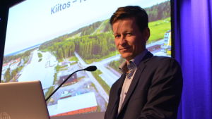 Dan Mollgren. Man står på scen framför ett podium med en dator. Han tittar in i kameran.