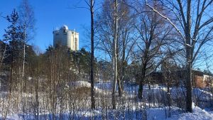Vattentornet uppe på backen. Överst syns också observatoriet som en liten kupol. Ett vattentorn syns långt borta uppe på en backe. Vinterlandskap, solsken. Buskage och byggnader syns också i förgrunden.