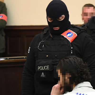 Den huvudmisstänkta Salah Abdeslam (till höger) sitter omringad av  belgiska specialstyrkor i rättegångssalen strax innan rättegången ska inledas. 