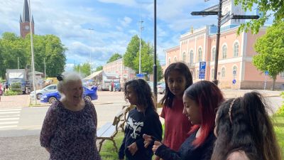 En äldre kvinna och sex flickor vid en bänk invid torget i Lovisa. De ser på varandra, talar och ler.